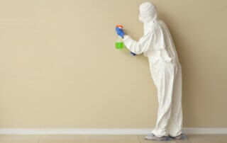 Technician in a protective suit and mask spraying a bare wall during a meth clean up